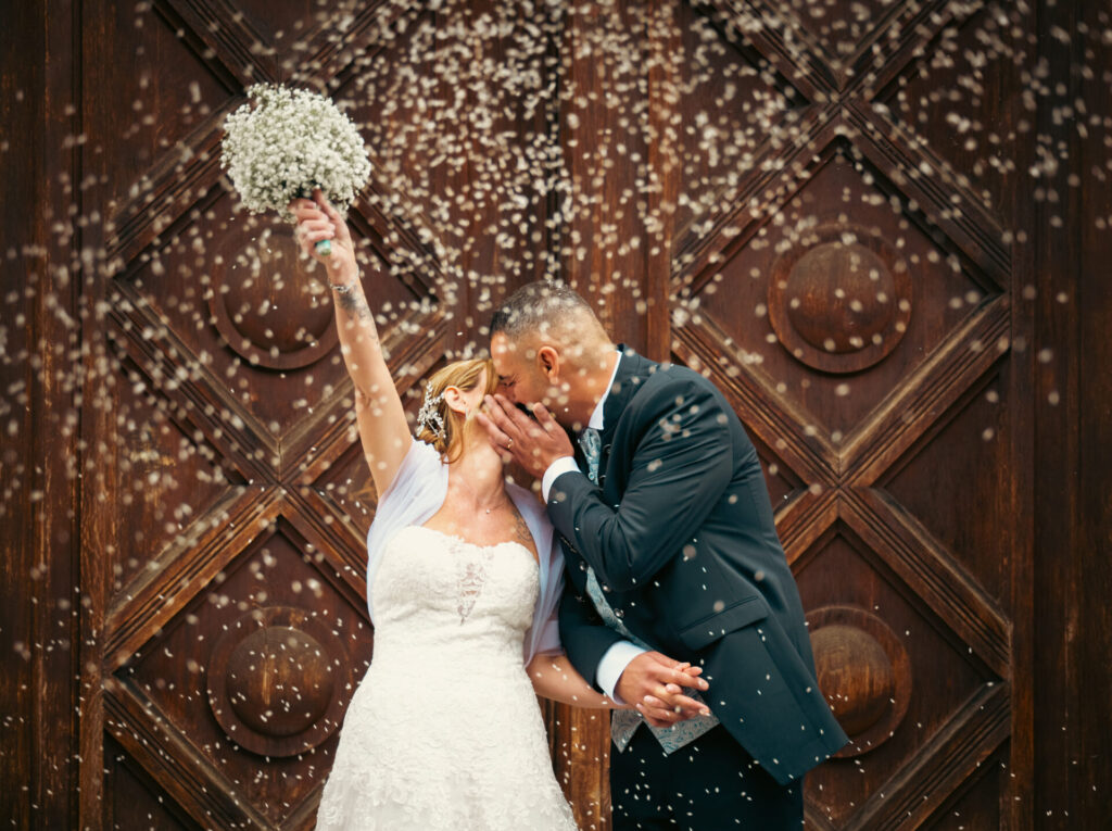 Coppia di sposi che si bacia fuori dalla chiesa mentre vengono lanciati chicchi di riso, con la sposa che solleva il bouquet al cielo