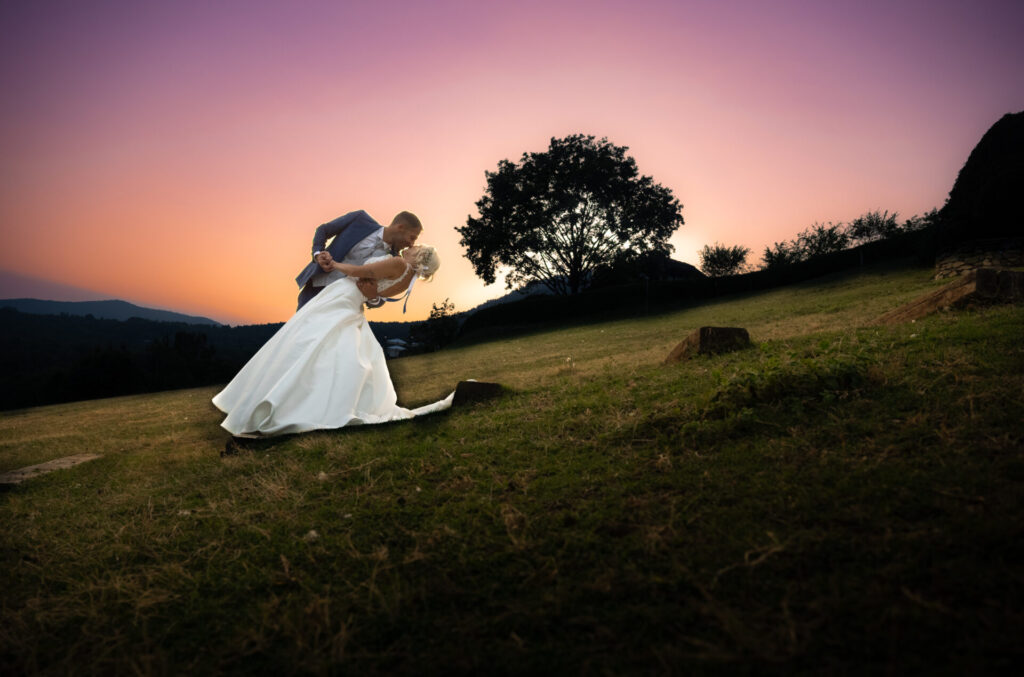 Coppia ripresa intera a colori con sfumature del tramonto sullo sfondo, lui che fa il casquè con la sposa inclinati in un momento romantico