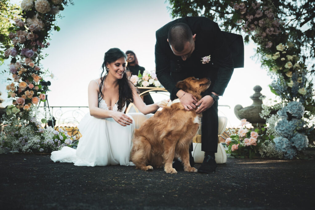 Coppia durante la cerimonia, con un arco di fiori sullo sfondo, la sposa accovacciata accarezza il cane mentre lo sposo si china per coccolarlo