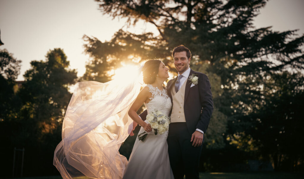Coppia immersa nella natura con il sole che illumina il velo della sposa, lei guarda lui con amore mentre lui guarda in camera