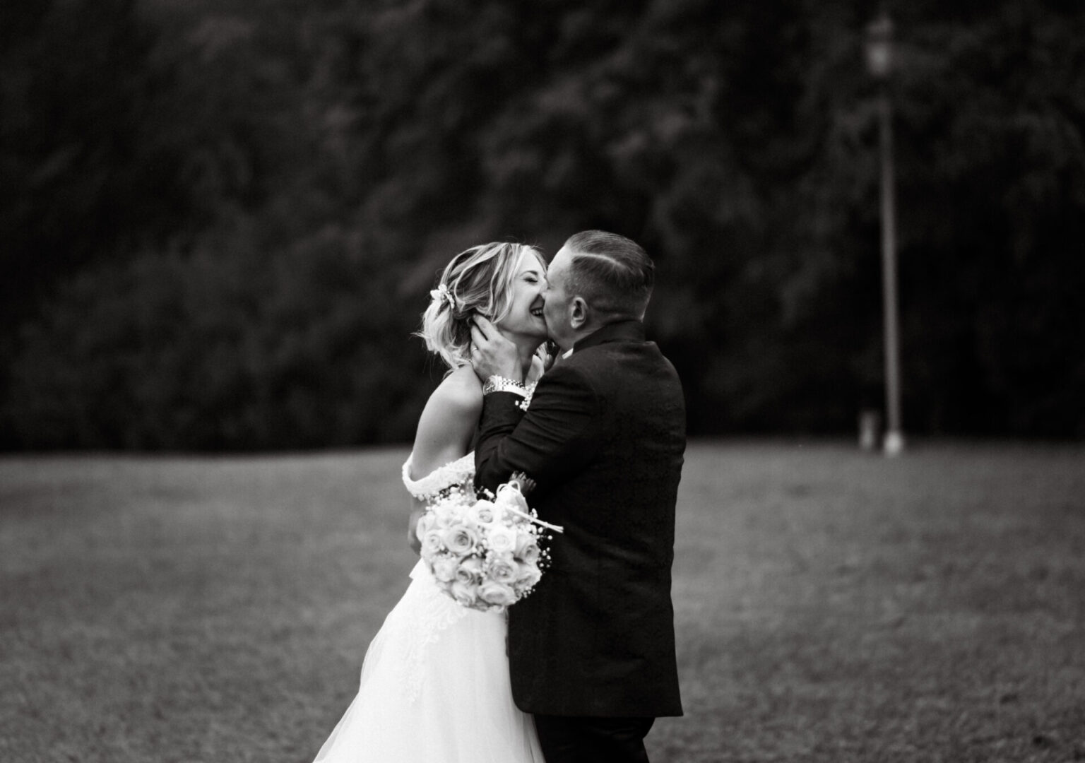 Foto in bianco e nero di una coppia che si bacia appassionatamente durante il matrimonio, immersa nella natura, con la sposa che sorride